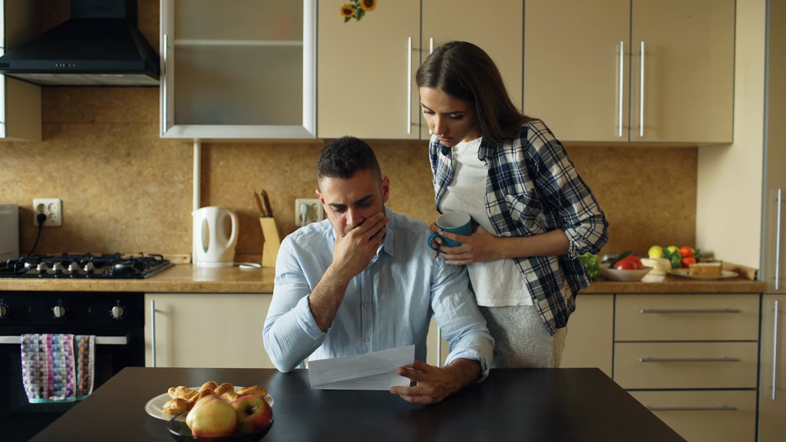 Un homme lit un courrier d'un air préoccupé, tandis qu'une femme l'entoure de ses bras dans une cuisine, tôt le matin.